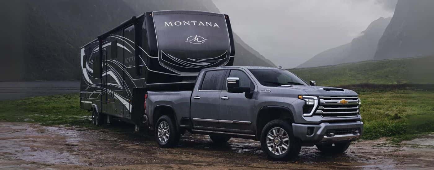 A gray 2024 Chevy Silverado 2500 HD High Country is shown towing a trailer on muddy ground.