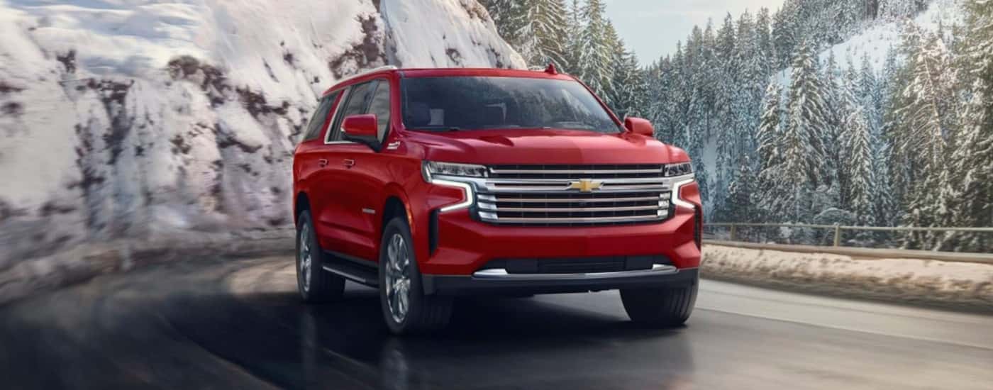 A red 2023 Chevy Suburban High Country is shown driving past snow-covered trees.