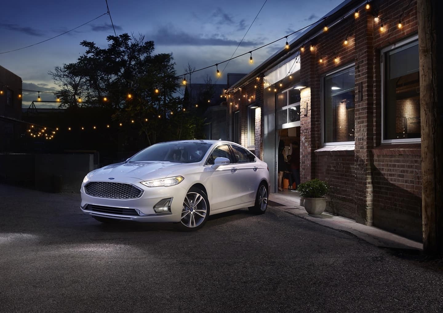Used Ford Fusion Titanium in white in front of a red brick building with patio string lights above.