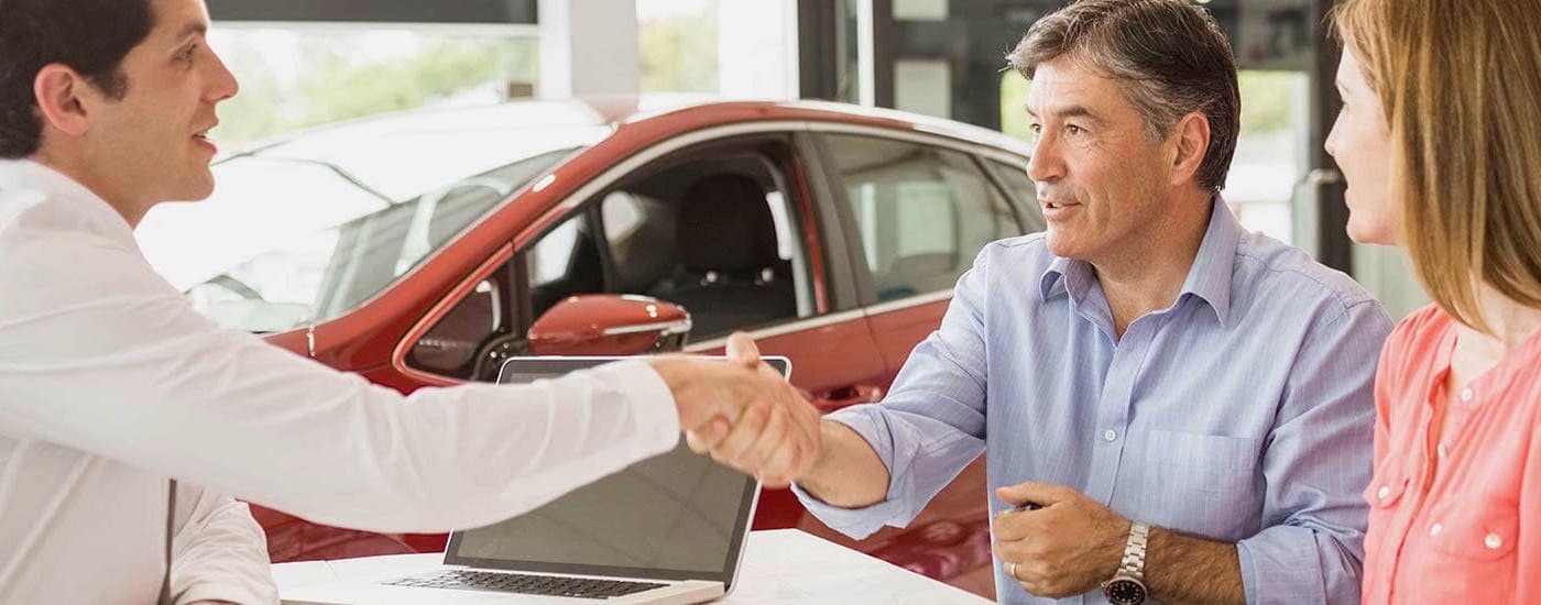 The salesman at your preferred use car dealer, Blossom Chevrolet, is shaking the hands of a new customer in a dealership showroom.