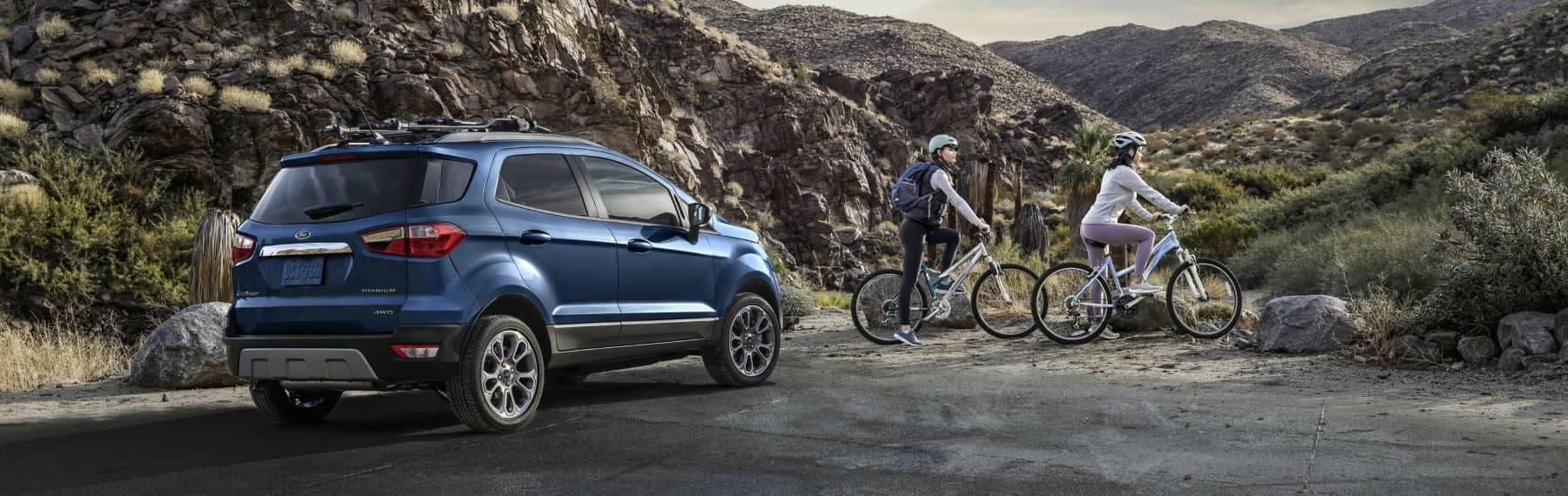 Two women on mountain bikes next to a parked Ford are preparing to ride a mountain trail