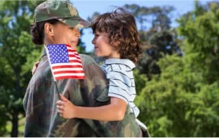 A soldier in uniform is holding a small child in her arms who is holding a small American flag.