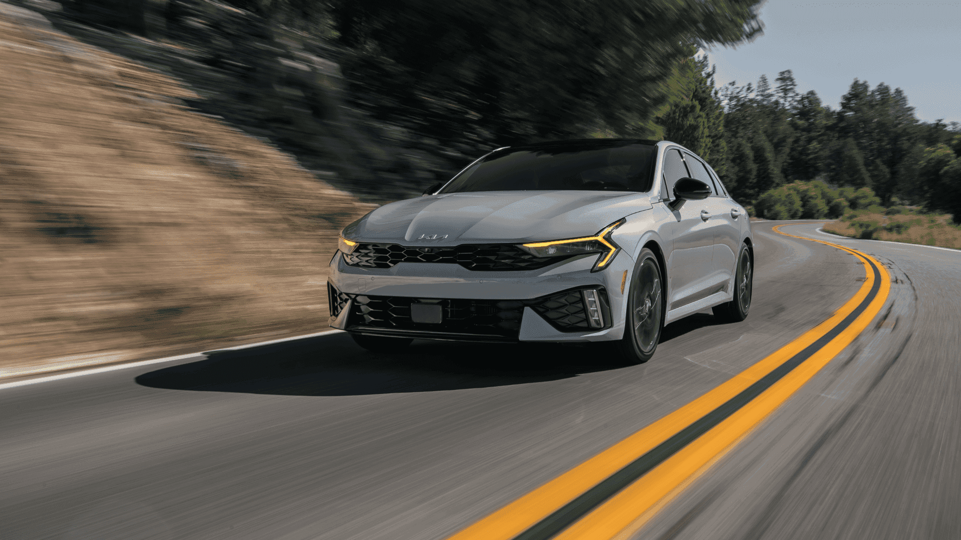 2025 Kia K5 midsize sedan driving through a curvy road, showcasing aggressive styling, LED headlights, and dynamic performance.