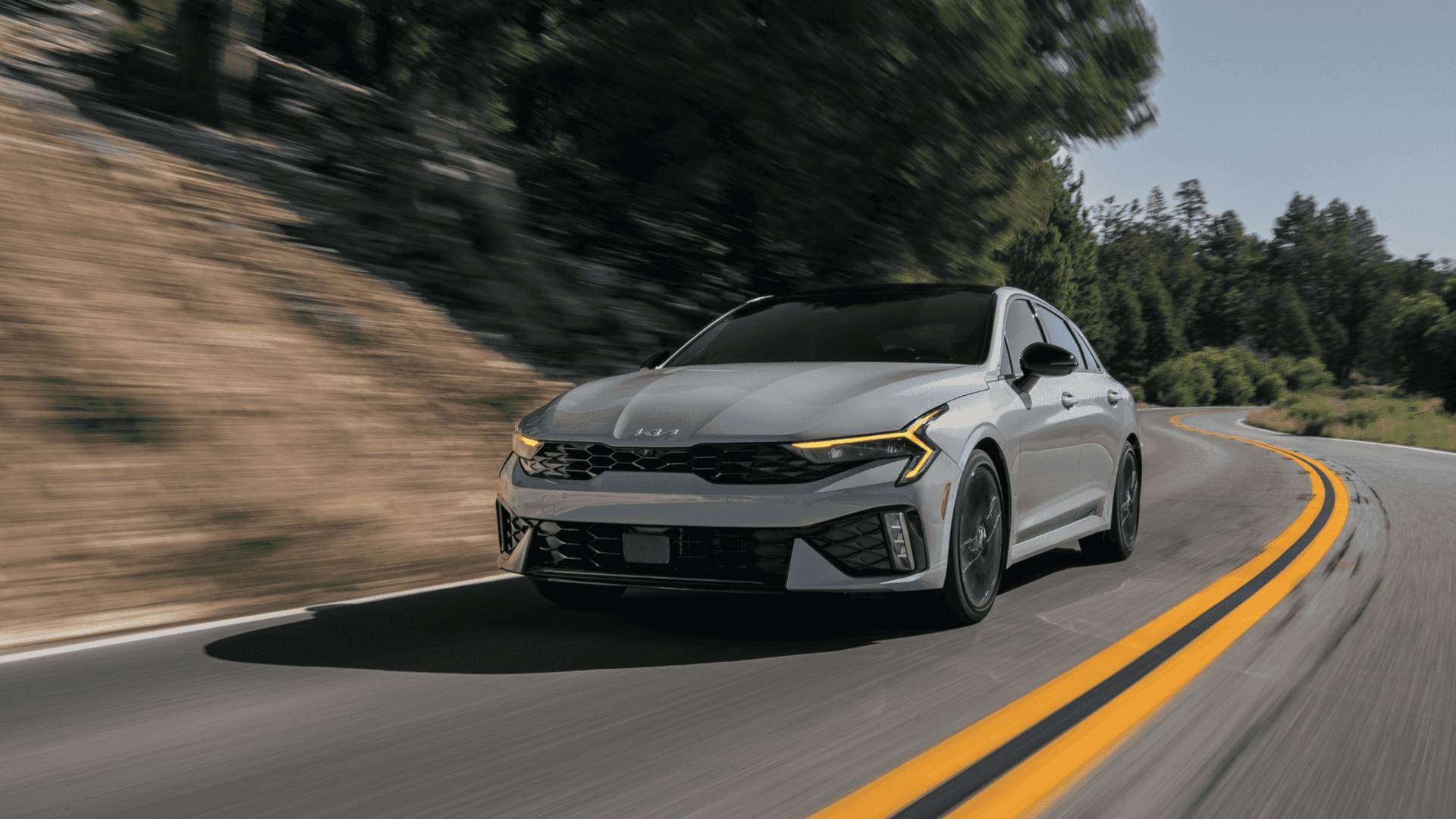 2025 Kia K5 midsize sedan cruising on a curvy road, boasting sporty performance and aggressive front styling.