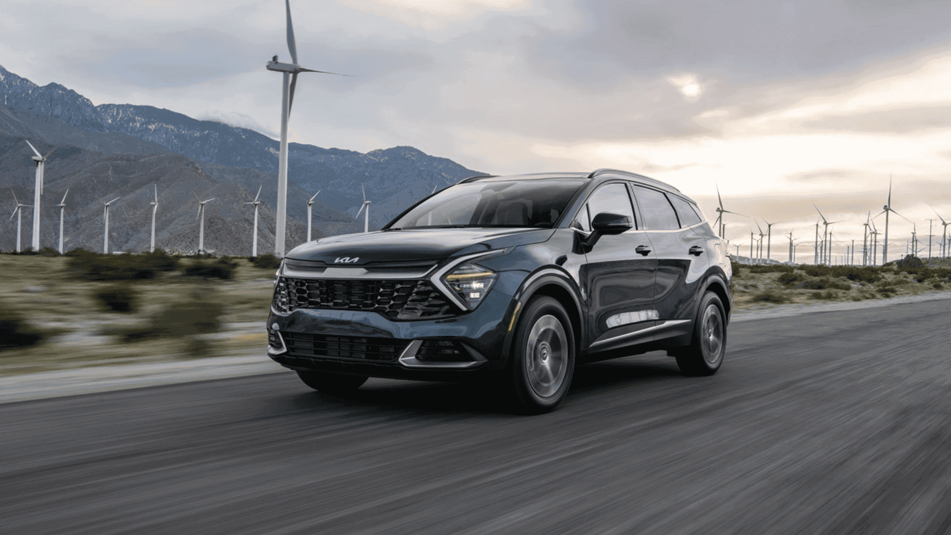2025 Kia Sportage Hybrid SUV driving past wind turbines, showcasing its fuel-efficient hybrid powertrain and bold exterior styling.