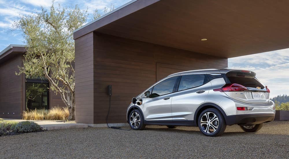 A silver 2020 Chevy Bolt is shown charging outside of a modern home.