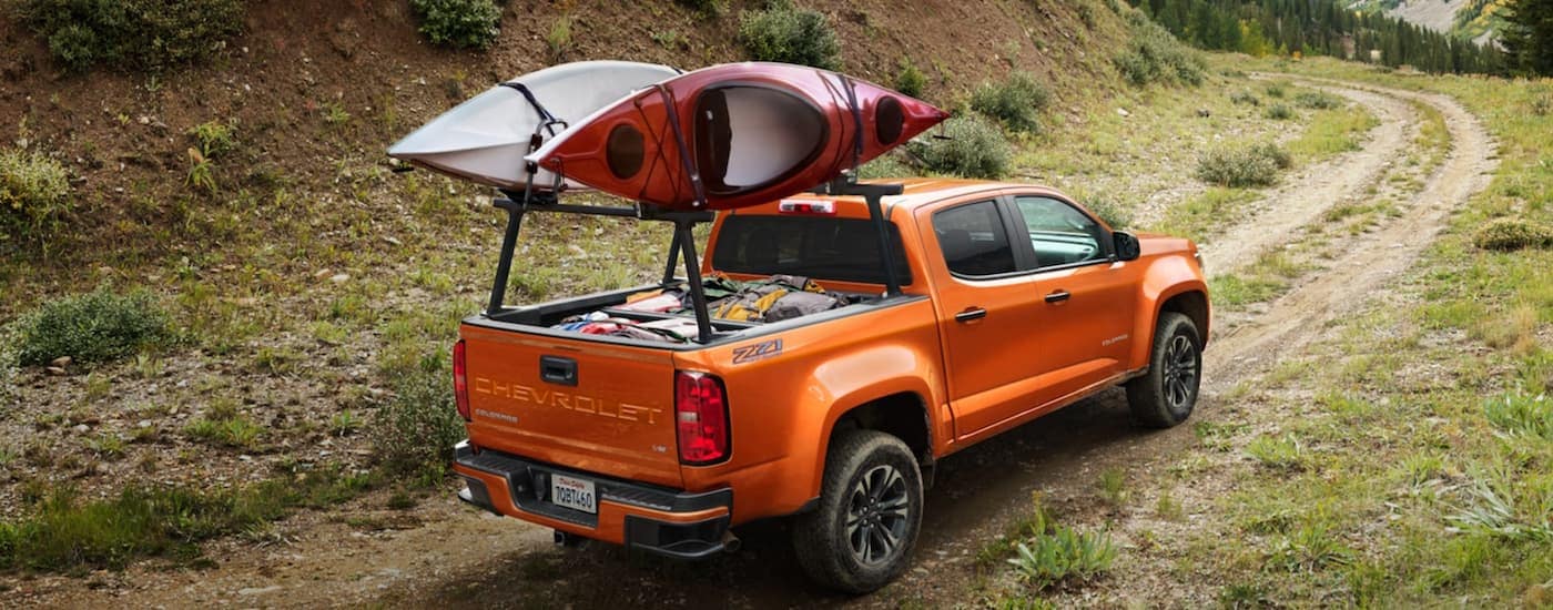 An orange 2022 Chevy Colorado Z71 is shown driving on a dirt road with kayaks mounted on bed racks.