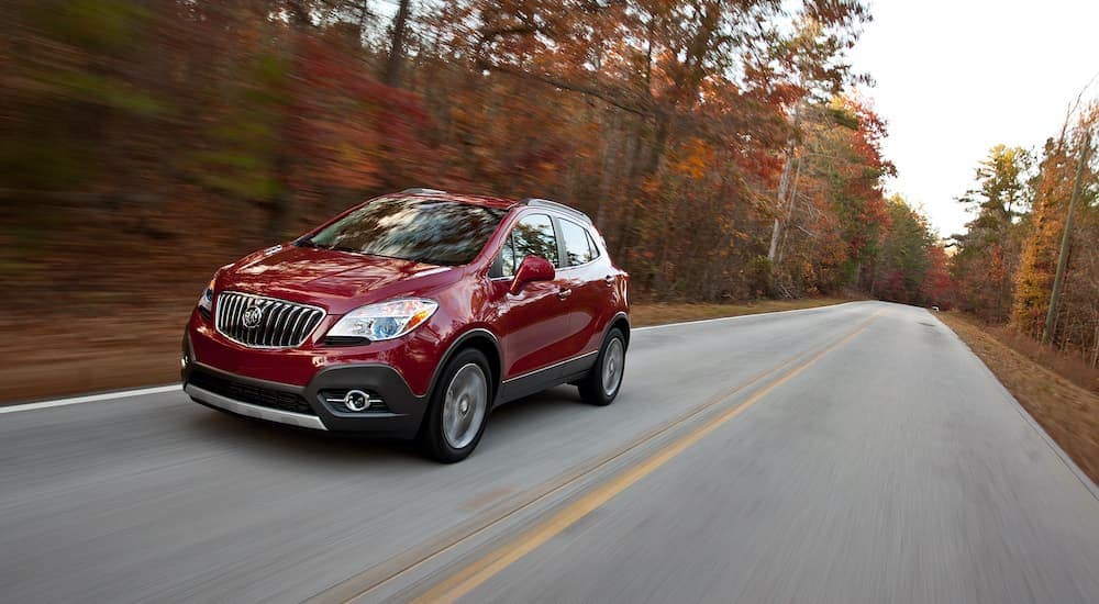 A red 2013 Buick Encore is shown driving down a tree lined road.