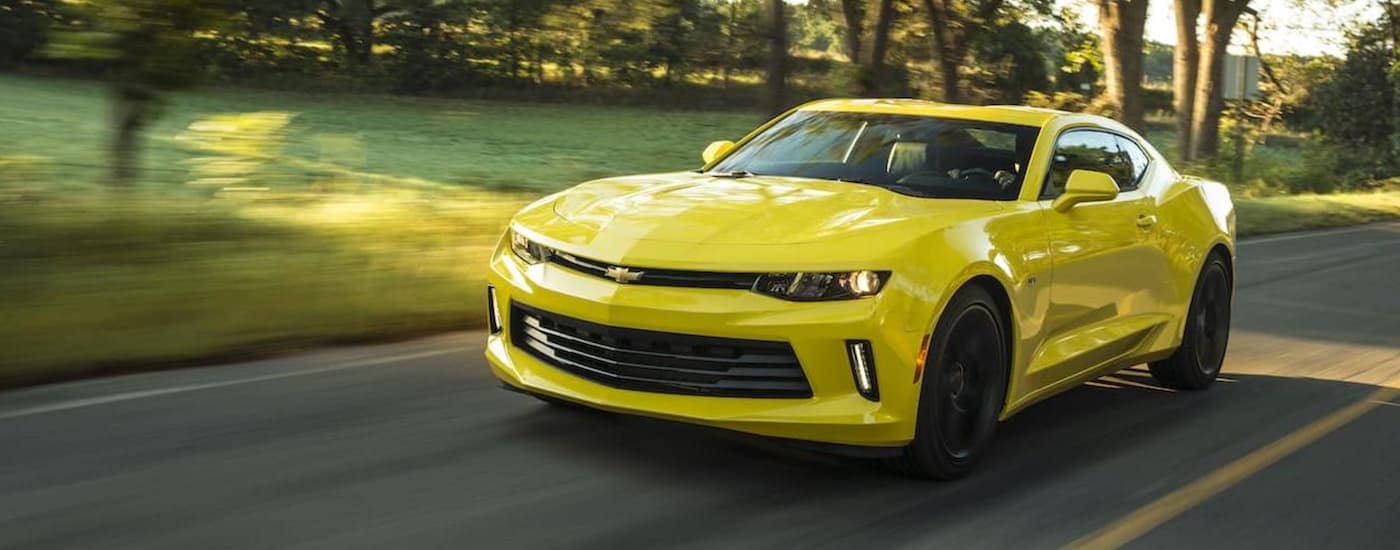 A yellow 2018 Chevy Camaro is shown driving down a tree lined road.