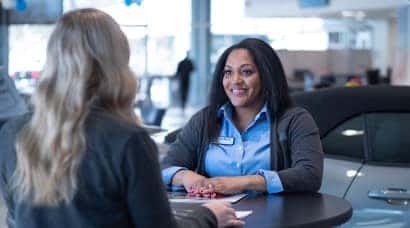 A woman sitting at a table inside a car dealership talking to another woman.