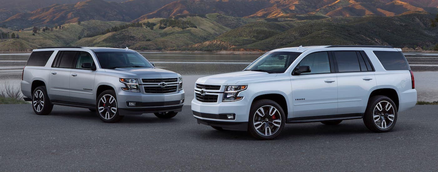 A silver 2020 Chevy Suburban and a white 2020 Chevy Tahoe RST are parked in front of a pond and mountains.