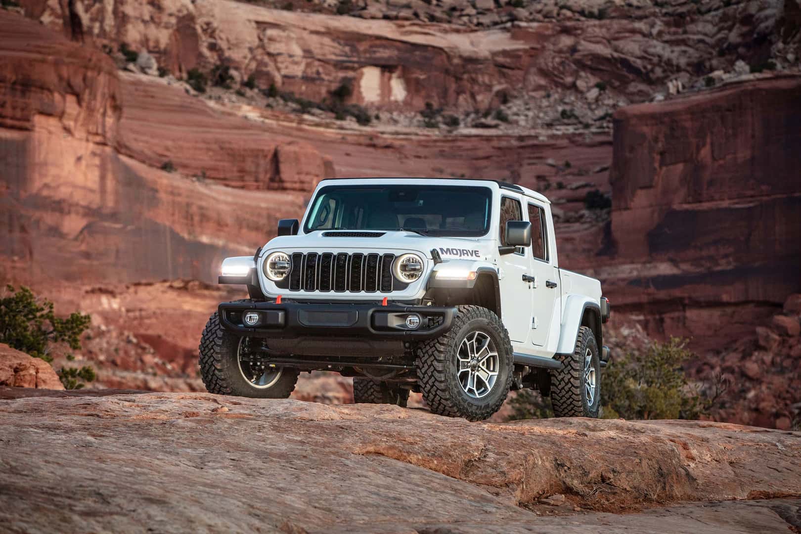 A white Jeep Gladiator Mojave pickup truck parked on red rocky terrain with a canyon in the background.