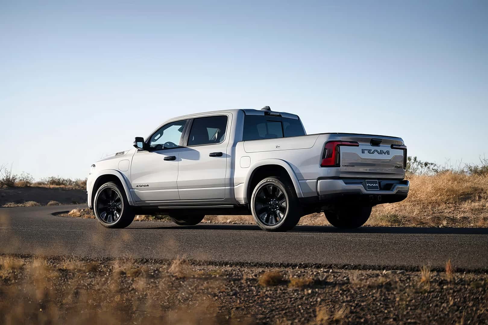 A gray Ram 1500 REV electric truck is shown from a rear three-quarter view, driving on a winding paved road through a desert landscape. The truck has black wheels and a lighted RAM logo on the tailgate. The sun is setting behind a distant hill.