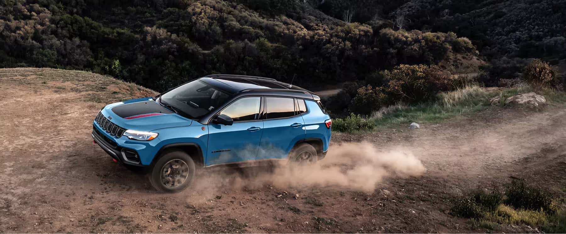 2025-Jeep-Compass-driving-along-a-dirt-hill-by-a-forest-valley