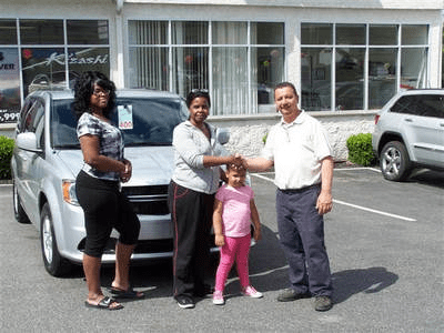 A family of three standing in front of their vehicle shaking hands with an employee from the dealership