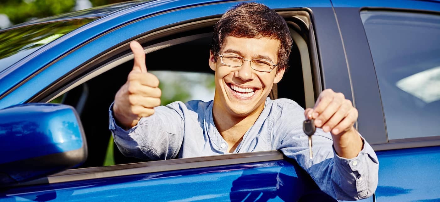 Close up of young man smiling holding car keys, giving a thumbs up sign out a car window