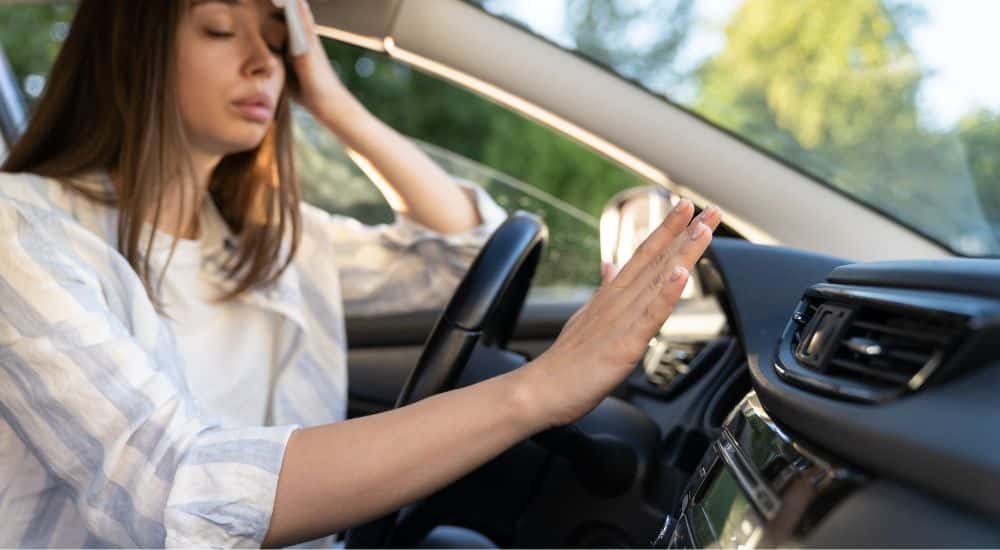 A woman dabbing sweat from her forehead in a car with faulty AC.