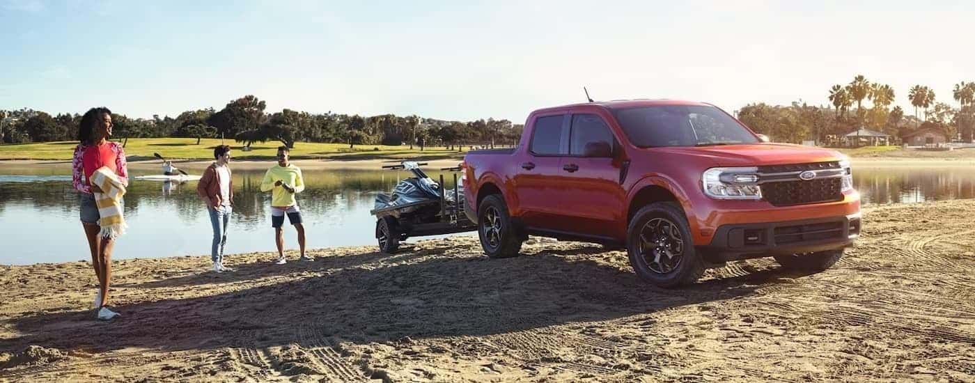 A red 2023 Ford Maverick is shown parked on a beach hooked up to a jet ski trailer.