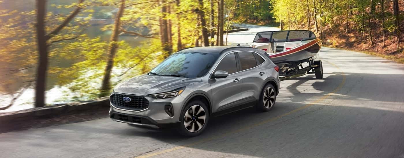 A silver 2024 Ford Escape is shown towing a boat on a winding forest road.