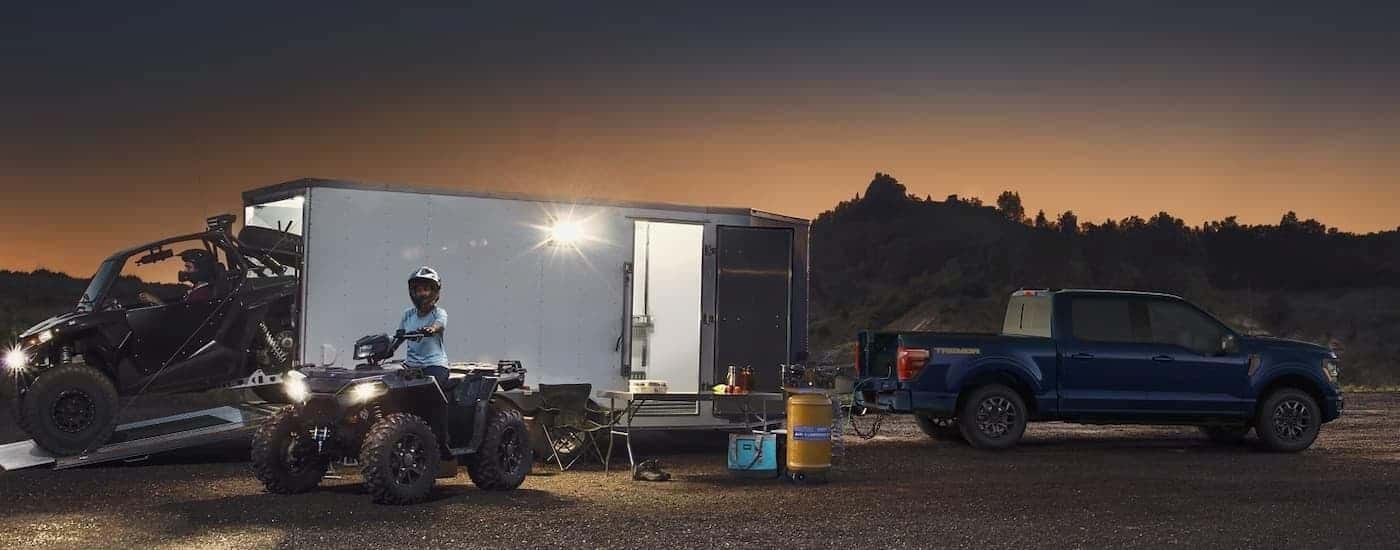 A blue 2025 Ford F-150 Tremor is parked near an enclosed trailer full of ATVs.