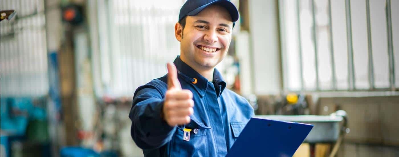 A vehicle mechanic is shown giving a thumbs up at the camera inside of a repair shop.