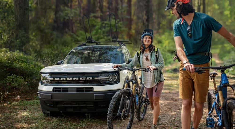 A couple walking their bikes away from a white 2024 Ford Bronco Sport Outer Banks parked in the woods.