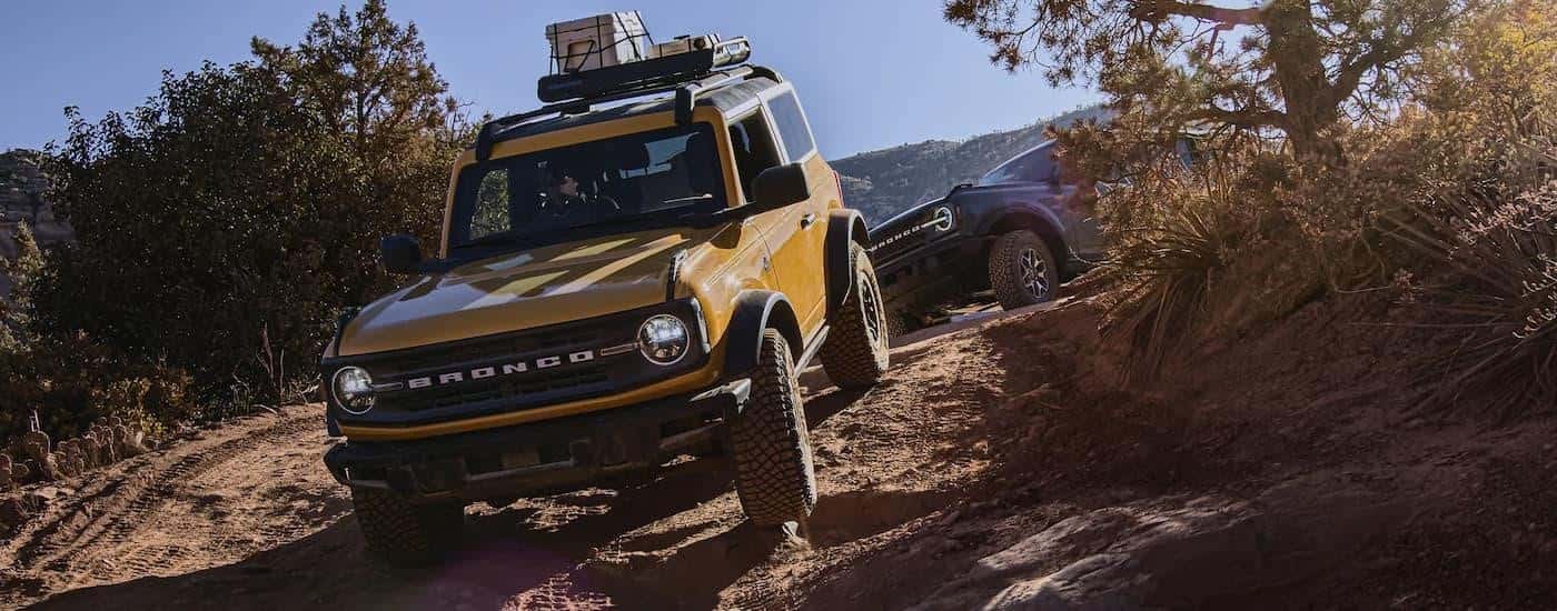 A yellow and a grey 2022 Ford Bronco are shown off-roading on a steep dirt trail.