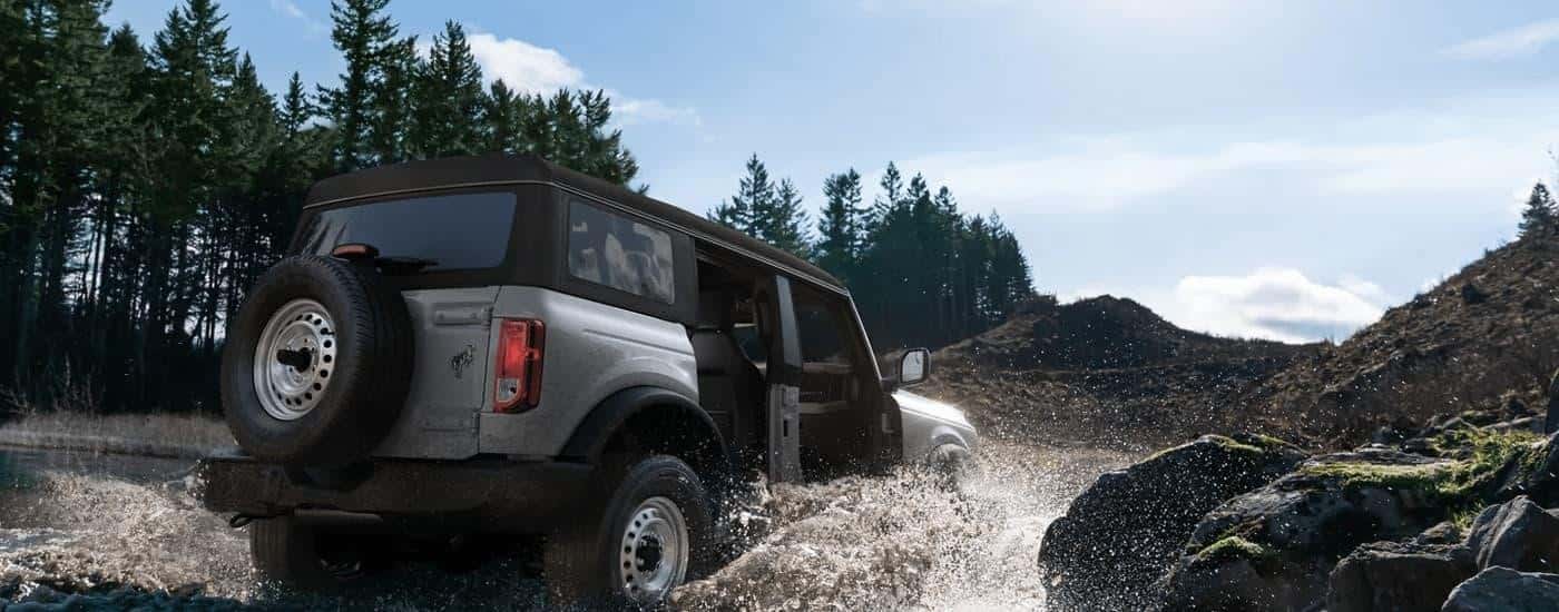 A silver 2023 Ford Bronco for sale is shown driving through a river.