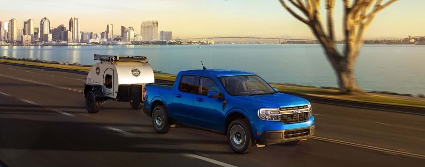 A blue 2023 Ford Maverick is shown towing a small silver camper away from a city.