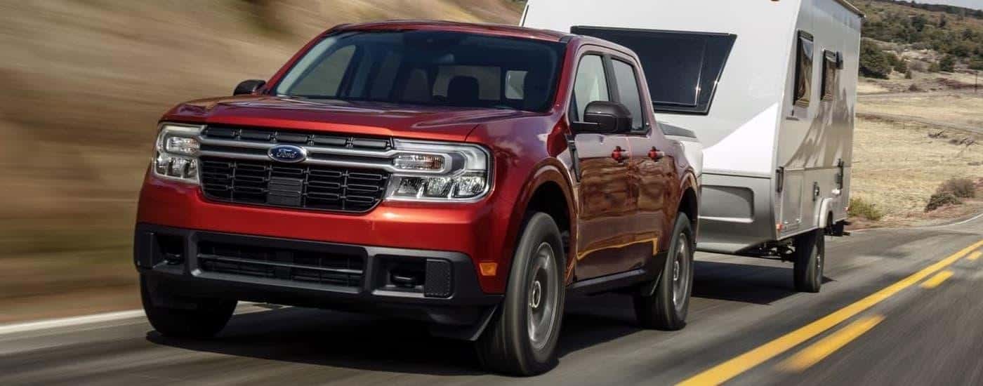 A red 2024 Ford Maverick Lariat is shown towing a camper on an open highway.