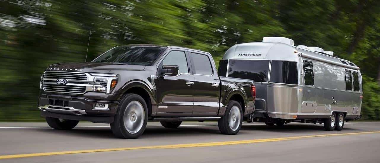 A grey 2024 Ford F-150 Platinum is shown towing a silver camper trailer on a tree-lined road.