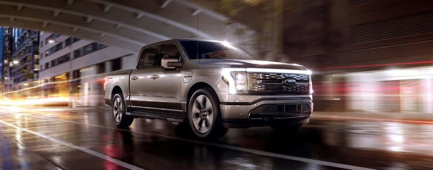 A silver A silver 2023 Ford F-150 Lightning is shown driving under a city bridge at night.