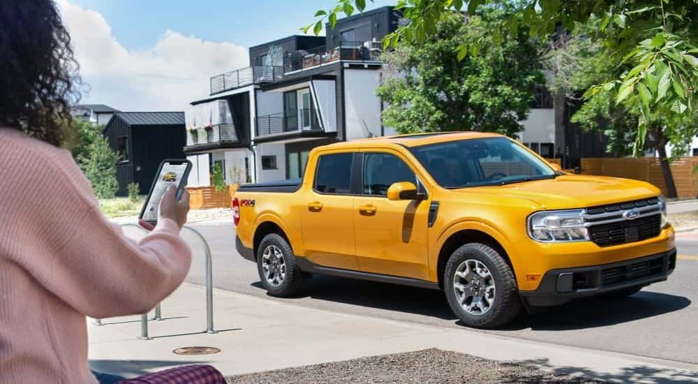 A woman using her smartphone to start a yellow 2024 Ford Maverick parked on a street.