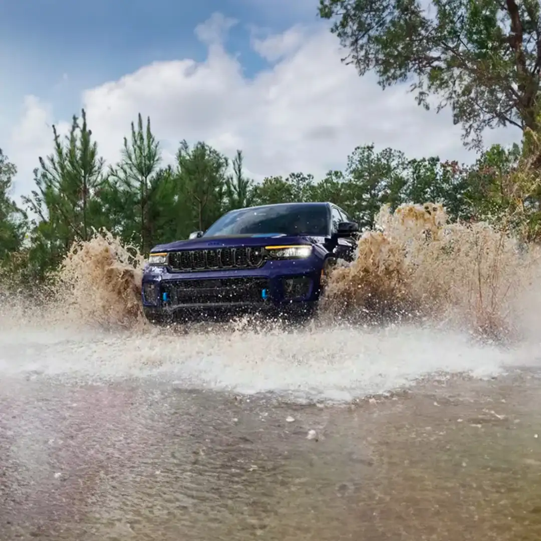 A blue 2024 Jeep Grand Cherokee Trailhawk 4xe fording through a muddy stream, with water splashing to the windows.