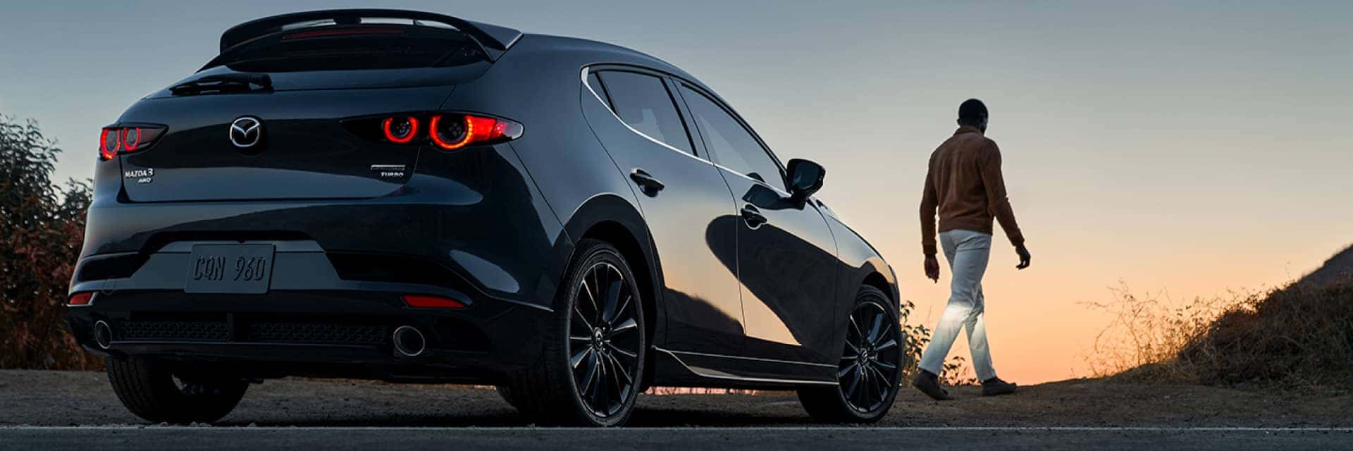 Rear view of a 2024 Mazda3 Hatchback at sunset with a person walking away from the vehicle