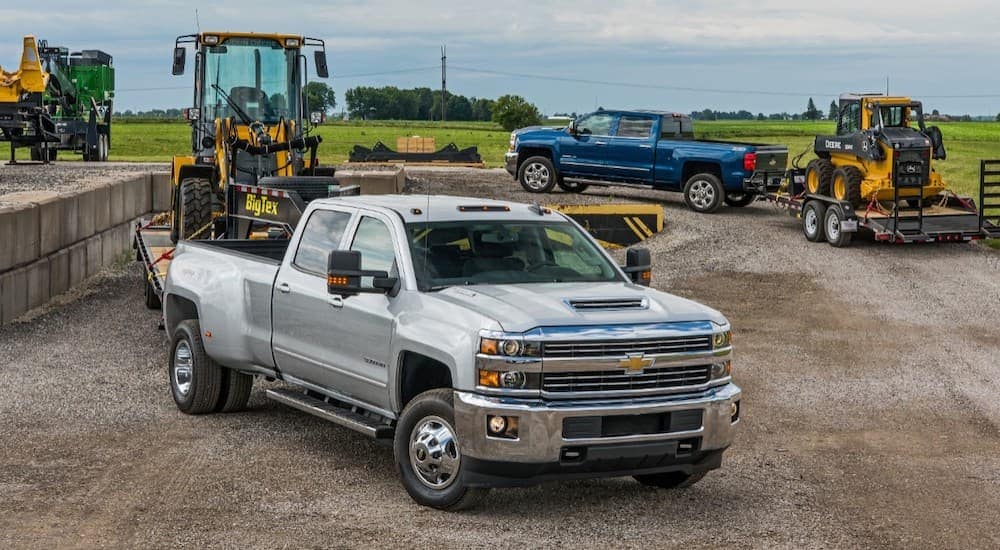 A silver and a blue 2018 Chevy Silverado 3500HD are hooked up to trailers with machinery.