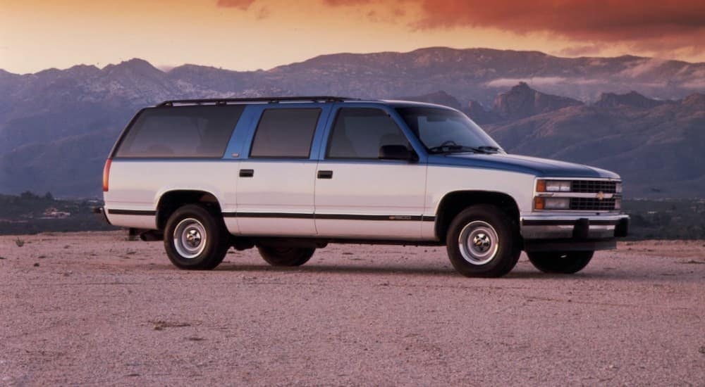 A blue and white 1992 Chevy Suburban, one of the older Chevy SUVs, is parked in front of mountains at sunset.
