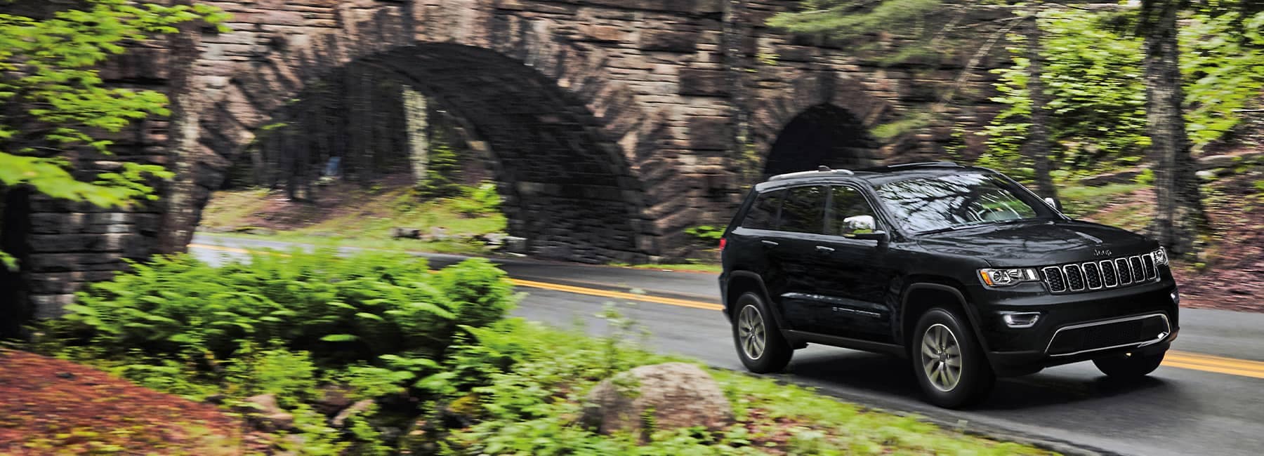 2022 Jeep Grand Cherokee WK Limited driving on forest lined road, with stone bridge behind