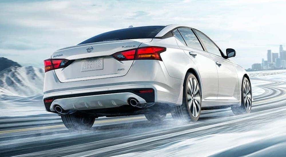 A white 2021 Nissan Altima is shown from the rear driving on a snowy road towards a city.