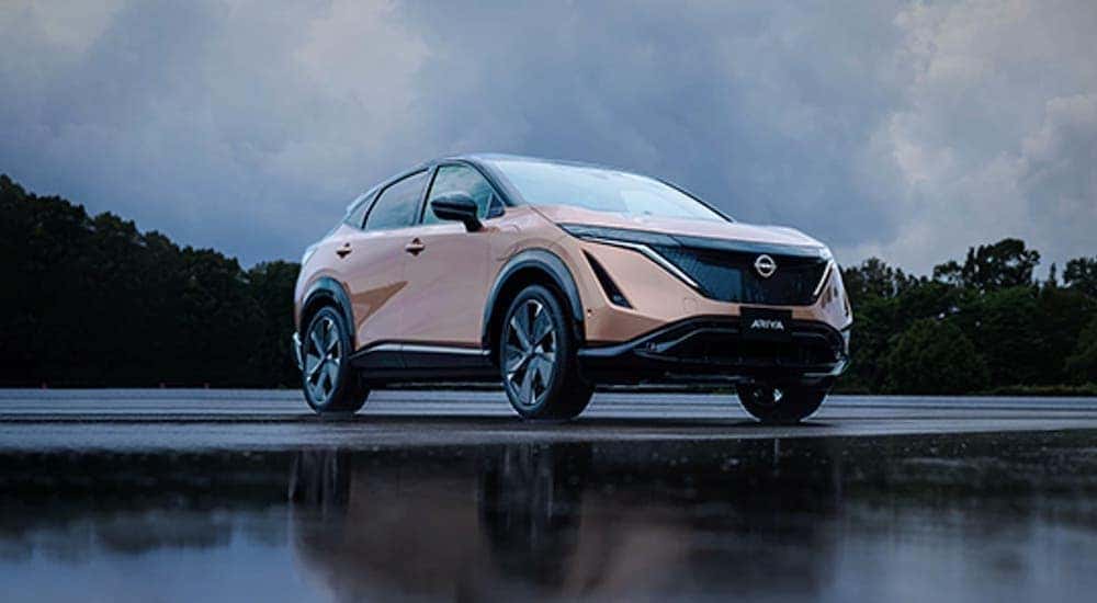 A pale orange 2021 Nissan Ariya is parked on a wet lot in front of trees near a Gallatin Nissan dealership.