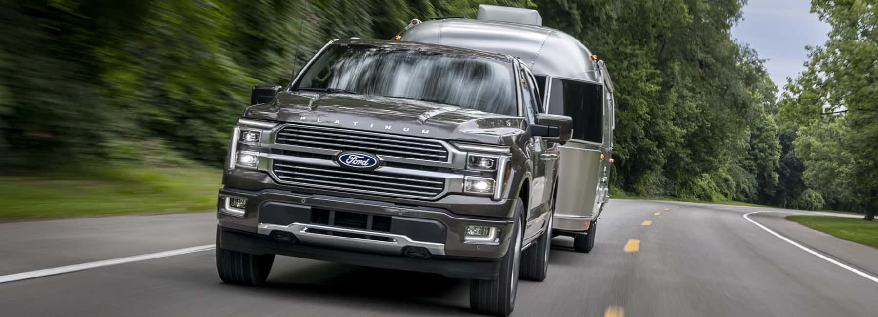 2025-Ford-F-150-pulling-a-silver-trailer-through-a-wooded-highway