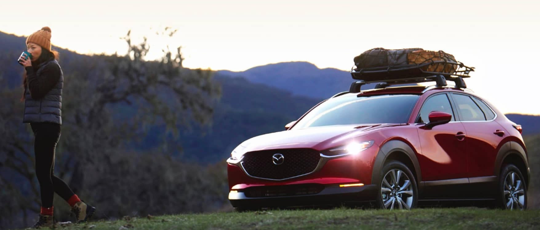 Red Mazda CX-30 parked on a grassy knoll in-front of a mountain range with a woman drinking coffee