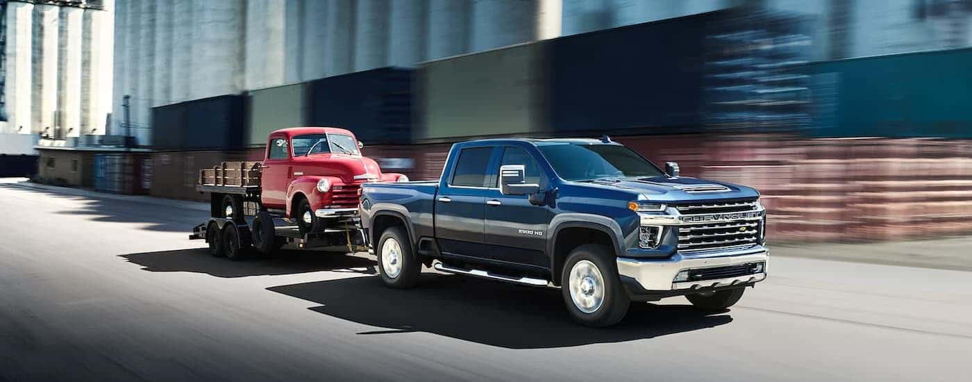 A blue 2022 Chevy Silverado 2500HD is shown towing a red truck down a city street