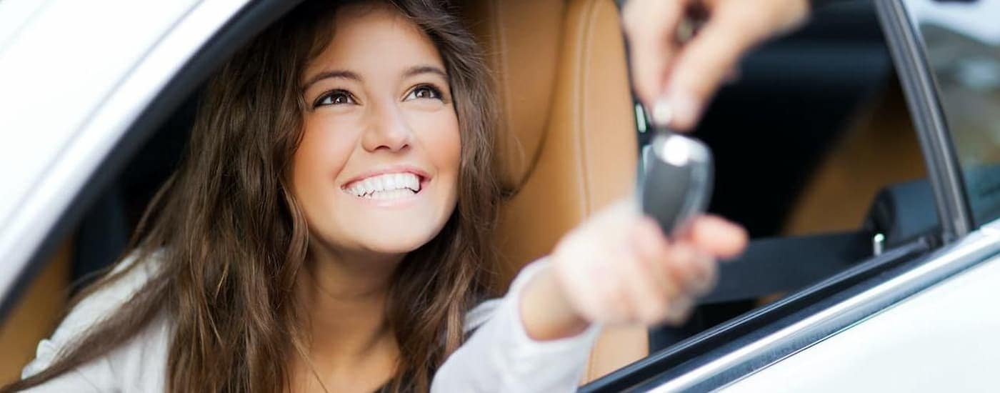 A woman is smiling from the front seat of a car as a salesperson passes a car key through the window.