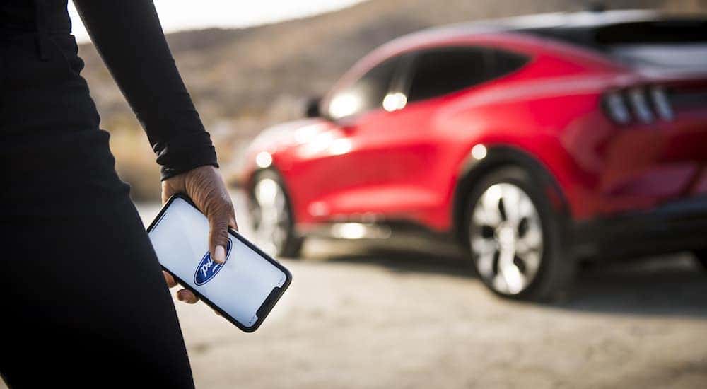 A close up shows a person holding a phone with the FordPass Connect app in front of a red 2022 Ford Mustang-Mach-E.