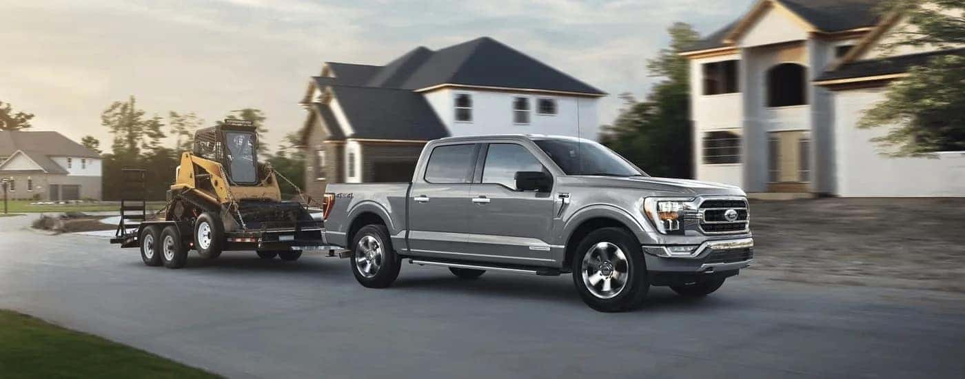 A silver 2023 Ford F-150 is shown towing heavy machinery through a road lined with houses.