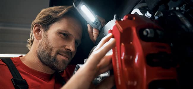 A Porsche certified technician using a flashlight to inspect brakes