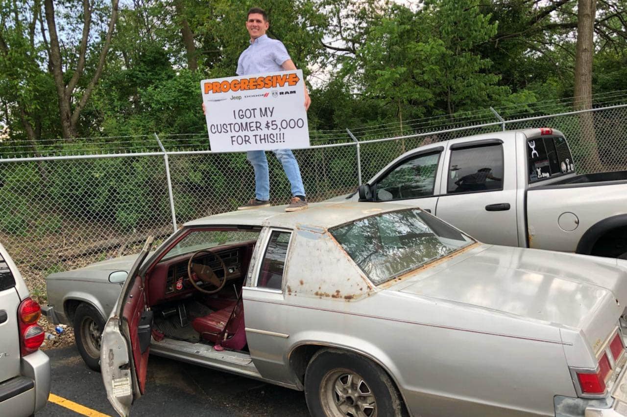 smiling Progressive customer standing on top of the trade-in car they received $5000 for