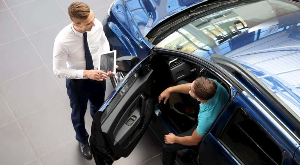A salesman is showing a blue used car to a buyer who is sitting in the driver's seat.