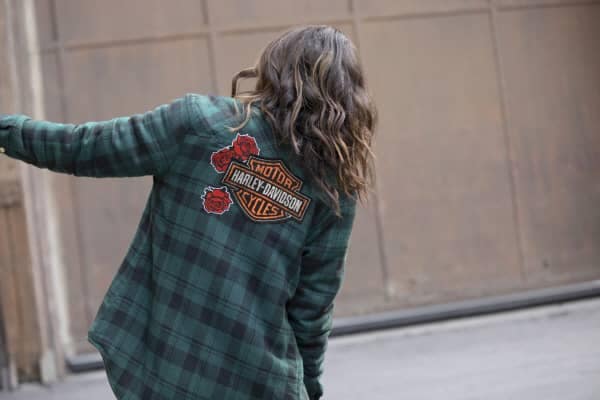 A women facing away with long, brown, wavy hair leans to the right wearing a green plaid Harley-Davidson long sleeve flannel shirt from the Harley-Davidson Garage collection.